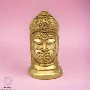 May include: A gold-colored metal sculpture of a deity's head with closed eyes and a mustache. The sculpture is detailed with intricate carvings and has a crown-like headdress.