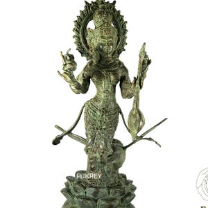 Może przedstawiać: Szczegółowa, brązowa statua bóstwa, stojąca na podstawie z kwiatu lotosu. Postać ma wiele ramion i jest ozdobiona skomplikowanymi detalami, w tym aureolą. Statua jest przedstawieniem postaci religijnej.