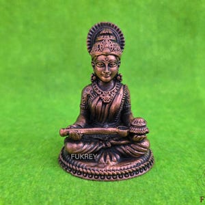 May include: A bronze-colored statue of a seated deity with ornate headdress and jewelry. The figure holds a musical instrument and a small pot. The base is detailed with a rope-like design. The word "FUKREY" is visible on the base.