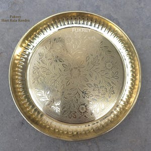 May include: A round, gold-colored metal plate with an intricate floral pattern etched into the surface. The plate has a decorative, ridged edge and the word "Fukrey" is visible. The plate is likely used for serving or display.