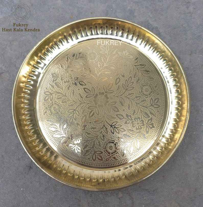 May include: A round, gold-colored metal plate with an intricate floral pattern etched into the surface. The plate has a decorative, ridged edge and the word "Fukrey" is visible. The plate is likely used for serving or display.