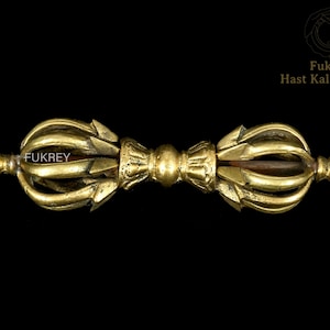 May include: A gold-coloured Vajra, a ritual object, against a black background. The Vajra has a central sphere, two symmetrical ends with ornate, curved designs, and pointed tips. The word "Fukrey" is visible on the object.
