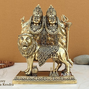 Puede incluir: Estatua de metal dorado de dos diosas montando un león. Las diosas llevan ropa y joyas adornadas. El león está de pie sobre una base con detalles intrincados. La estatua es un icono religioso hindú tradicional.