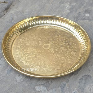 May include: A round, gold-colored brass plate with an embossed floral design in the center. The rim has a textured, ridged pattern. The plate is likely used for serving or display. The plate is approximately 10 inches in diameter.
