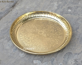 Puede incluir: Una placa redonda de latón dorado con un diseño floral en relieve en el centro. El borde tiene un patrón texturizado y acanalado. La placa probablemente se utiliza para servir o exhibir. La placa tiene aproximadamente 25 cm de diámetro.