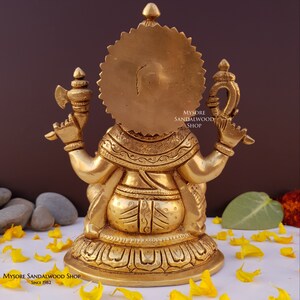 K&ouml;nnte beinhalten: Eine goldfarbene Messingstatue von Ganesha, dem hinduistischen Gott der Weisheit und des Wohlstands, der in meditativer Haltung sitzt. Die Statue ist aufwendig detailliert und hat einen Heiligenschein hinter Ganeshas Kopf. Die Statue sitzt auf einem Sockel mit einem Lotus-Design. Der Text "Mysore Sandalwood Shop Since 1982" ist auf dem Sockel sichtbar.