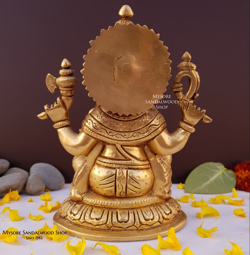 K&ouml;nnte beinhalten: Eine goldfarbene Messingstatue von Ganesha, dem hinduistischen Gott der Weisheit und des Wohlstands, der in meditativer Haltung sitzt. Die Statue ist aufwendig detailliert und hat einen Heiligenschein hinter Ganeshas Kopf. Die Statue sitzt auf einem Sockel mit einem Lotus-Design. Der Text "Mysore Sandalwood Shop Since 1982" ist auf dem Sockel sichtbar.