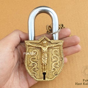 Puede incluir: Un candado de latón con un diseño detallado que presenta una figura de una persona con los brazos extendidos. El candado tiene un grillete plateado y está unido a una cadena.