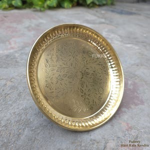 May include: A round, golden brass plate with an embossed floral design in the center. The rim of the plate has a textured, ridged pattern. The plate is sitting on a gray surface.