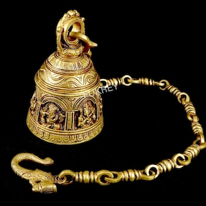 May include: A gold-coloured bell with detailed carvings and a chain. The bell has relief carvings of figures and patterns. The chain ends with a decorative hook. The bell is made of a shiny, metallic material.