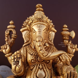 K&ouml;nnte beinhalten: Eine goldfarbene Metallstatue von Ganesha, dem hinduistischen Gott der Weisheit und des Wohlstands. Die Statue ist aufwendig detailliert und zeigt Ganesha mit seinem Elefantenkopf, R&uuml;ssel und vier Armen. Die Statue sitzt auf einem Sockel und tr&auml;gt eine kleine, verzierte Krone auf dem Kopf. Der Text "MYSORE SANDALWOOD SHOP" ist auf der rechten Seite der Statue sichtbar.