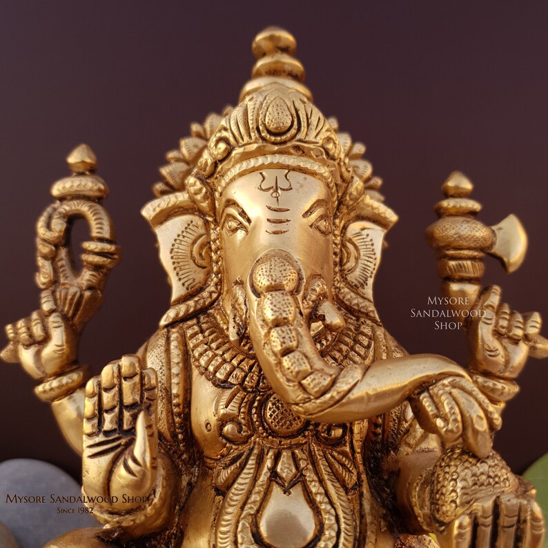 K&ouml;nnte beinhalten: Eine goldfarbene Metallstatue von Ganesha, dem hinduistischen Gott der Weisheit und des Wohlstands. Die Statue ist aufwendig detailliert und zeigt Ganesha mit seinem Elefantenkopf, R&uuml;ssel und vier Armen. Die Statue sitzt auf einem Sockel und tr&auml;gt eine kleine, verzierte Krone auf dem Kopf. Der Text "MYSORE SANDALWOOD SHOP" ist auf der rechten Seite der Statue sichtbar.