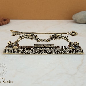 Op de afbeelding: Een gedetailleerde messing sculptuur van een boog en pijl, met ingewikkelde gravures en een decoratieve basis. De pijl is boven de boog geplaatst en de basis heeft tekst en de merknaam "Fukrey".