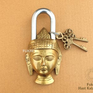 Puede incluir: Un candado dorado con un diseño de cabeza de Buda. El candado tiene un lazo de metal plateado y viene con dos llaves.