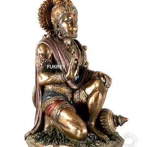 Puede incluir: Estatua de bronce de una deidad arrodillada, adornada con intrincadas joyas y un tocado. La figura tiene una expresión serena y está cubierta con una prenda morada y roja. La base de la estatua está detallada con follaje. El texto "Fukrey" es visible.