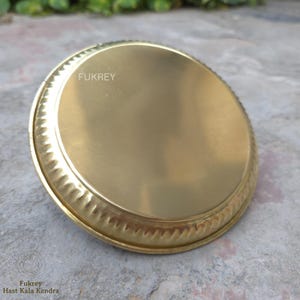 May include: A round, brass-colored plate with a decorative, ridged edge. The plate has a smooth, reflective surface and the word "FUKREY" is printed on the plate. The plate is sitting on a gray surface.