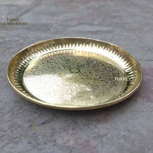 May include: A round, gold-colored brass plate with an intricate floral pattern etched into the center. The rim has a textured, ridged design. The plate is from Fukrey Hast Kala Kendra.