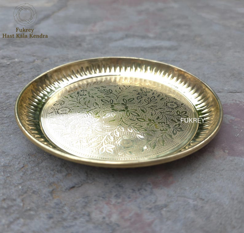 May include: A round, gold-colored brass plate with an intricate floral pattern etched into the center. The rim has a textured, ridged design. The plate is from Fukrey Hast Kala Kendra.