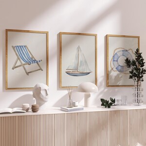 Puede incluir: Tres impresiones enmarcadas en acuarela con temática náutica. Las impresiones representan una silla de playa a rayas azules y blancas, un velero y un salvavidas azul y blanco. Los marcos son dorados y se exhiben en una pared blanca.