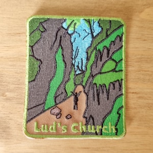 May include: Embroidered patch depicting a gorge scene with a figure. The patch has a green border and the text "Lud's Church". The design includes grey rock formations, green foliage, and a blue sky. Ideal for jackets, bags, or clothing.