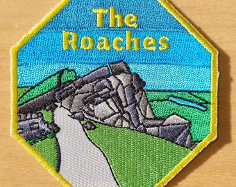 Könnte beinhalten: Ein gelber, achteckiger gestickter Aufnäher mit blauem Himmelhintergrund und grünem Grasvordergrund. Der Aufnäher zeigt eine graue Felsformation mit dem Text "The Roches" in gelben Buchstaben.
