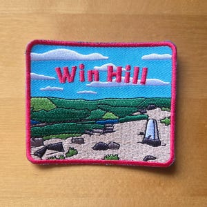 May include: A rectangular embroidered patch with a red border featuring a landscape scene of Win Hill. The design includes a bright blue sky with white clouds, green rolling hills, and a rocky foreground with a white stone trig point marker.