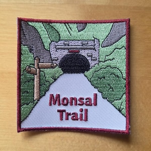 May include: A square embroidered patch featuring a stone tunnel entrance surrounded by green foliage and grey rock formations. A wooden signpost stands on the left. The text Monsal Trail is stitched in red on a light grey path leading to the tunnel.
