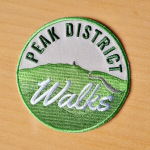 May include: Green and white embroidered patch with the text "Peak District Walks" in a white cursive font. The patch features a green hill with a winding path and a small building at the top.