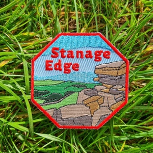 May include: A red octagon-shaped patch with the words "Stanage Edge" in red text. The patch features a colourful embroidered image of a rocky landscape.