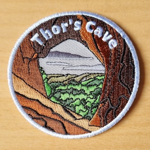 May include: Circular embroidered patch displaying "Thor's Cave" in white. The design shows a cave entrance, mountains, and green foliage. The patch has a light blue border and is a detailed landscape scene.