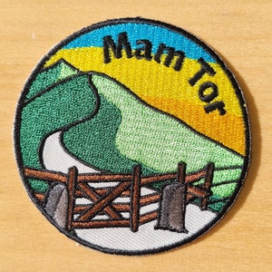 May include: A round embroidered patch featuring a green hill, a winding road, a wooden fence, and a sunset. The text "Manor" is written in a curved arc above the hill.