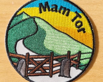 Könnte beinhalten: Ein runder gestickter Aufnäher mit einem grünen Hügel, einer kurvigen Straße, einem Holzzaun und einem Sonnenuntergang. Der Text "Manor" ist in einem Bogen über dem Hügel geschrieben.