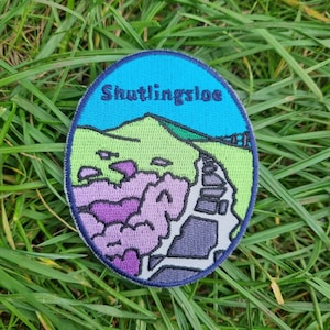 May include: An embroidered patch featuring the text "Shutlingsloe" and a depiction of a mountain and a winding path, in shades of blue, green and purple.