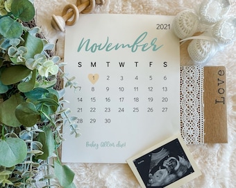 May include: A white calendar with the month of November highlighted in blue. The number 1 is circled with a pink heart. The text "Baby Allen due!" is written below the calendar. There is a sonogram image, a pair of white crocheted baby booties, a wooden sign that says "Love", and a wooden rattle in the image.