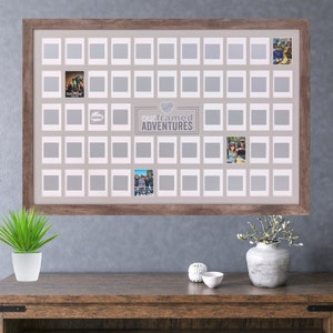 Könnte beinhalten: Ein brauner Holzrahmen mit weißem Hintergrund und 49 leeren Fotoplätzen. Der Rahmen trägt den Text "Our Framed Adventures" mit einem Herzsymbol in der Mitte. Der Rahmen hängt an einer grauen Wand.