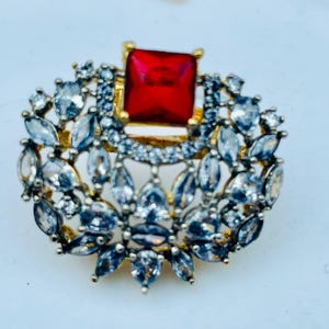May include: A gold-toned ring with a large red square-shaped stone surrounded by smaller clear stones.