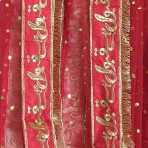 May include: Red sheer fabric with gold embroidered trim featuring Arabic calligraphy. The fabric has a dotted pattern.