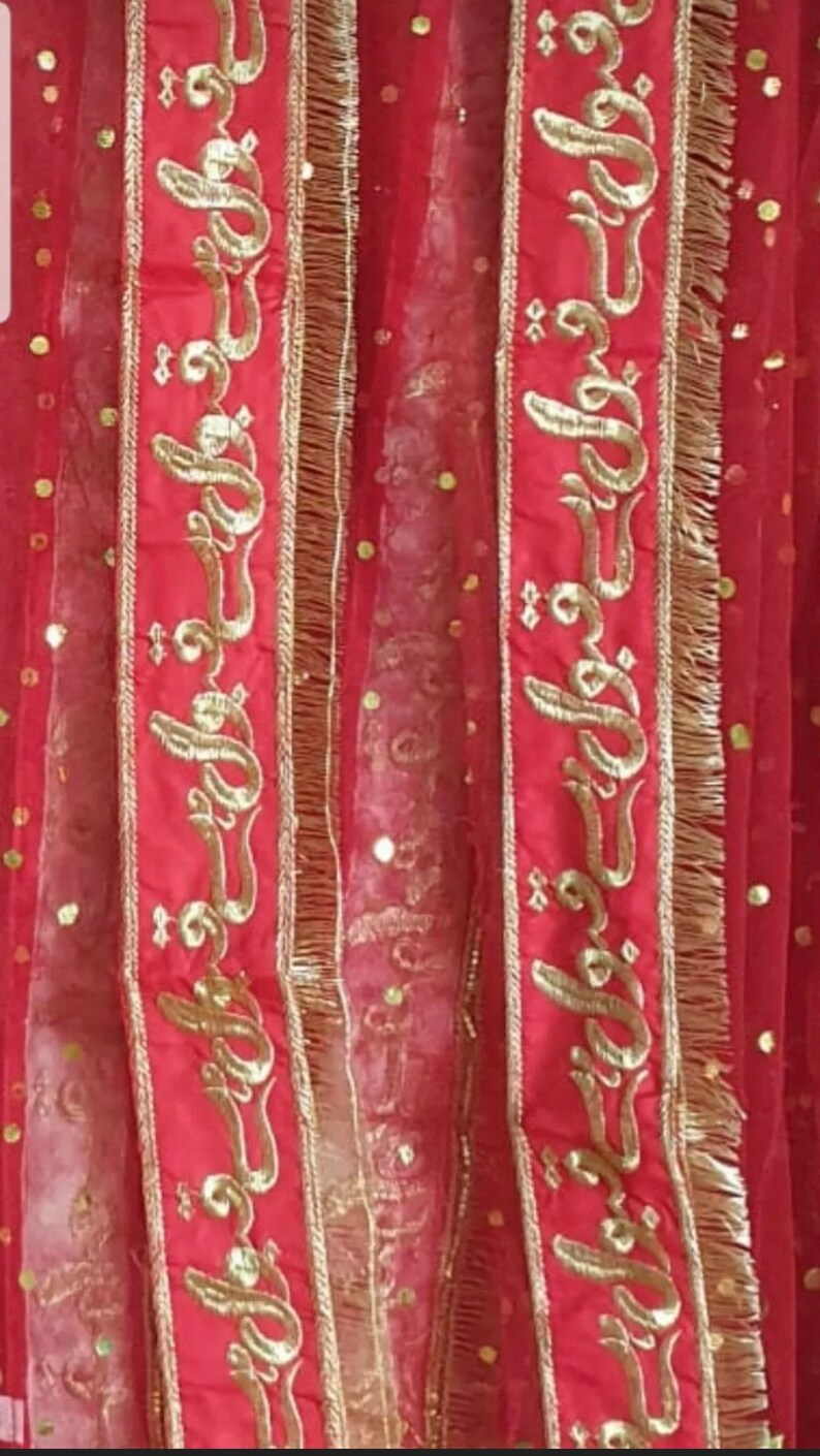 May include: Red sheer fabric with gold embroidered trim featuring Arabic calligraphy. The fabric has a dotted pattern.