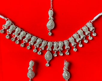 Könnte beinhalten: Ein silberfarbenes Schmuckset mit Halskette, Ohrringen und einem Haarschmuck, alle mit funkelnden, klaren Steinen besetzt. Halskette und Ohrringe haben ein tropfenförmiges Design. Das Set wird vor rotem Hintergrund präsentiert.