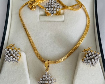 May include: A gold-toned jewelry set featuring a necklace, ring, and earrings, all adorned with sparkling, clear gemstones. The necklace has a textured chain, and the pendant, ring, and earrings share a diamond-shaped design. The set is displayed in a black and white jewelry box.
