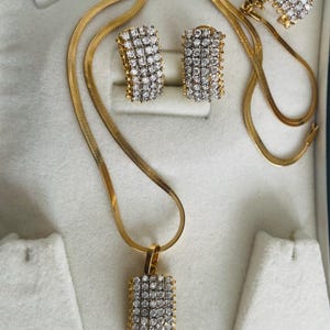 Peut inclure: Un ensemble de bijoux doré comprenant un collier, un pendentif et des boucles d'oreilles assorties. Le pendentif et les boucles d'oreilles ont un design rectangulaire, incrusté de petites pierres scintillantes. Le collier est une chaîne plate en chevrons.