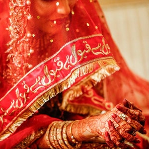 May include: A bride wearing a red veil with gold embroidery and a red dress with gold accents. Her hands are decorated with henna and she is wearing gold bangles.