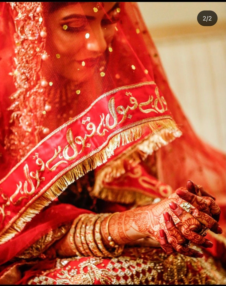 May include: A bride wearing a red veil with gold embroidery and a red dress with gold accents. Her hands are decorated with henna and she is wearing gold bangles.