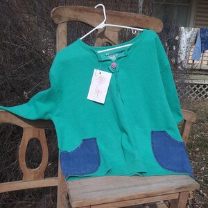 May include: A teal green shirt with a button and a tie at the neck. The shirt has two denim pockets on the bottom. The shirt is hanging on a wooden chair.
