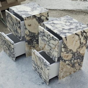 Puede incluir: Dos mesitas de noche con tapa de mármol y cajones abiertos. El mármol tiene un patrón crema y negro. Los cajones son blancos. Las mesitas de noche tienen forma de cubo y están sobre una superficie de hormigón. Losas de mármol adicionales visibles.