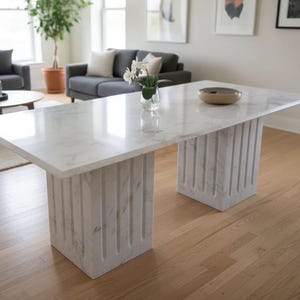 May include: A rectangular dining table featuring a smooth white marble top with grey veining. The table is supported by two substantial, rectangular pedestals made of matching white marble, each characterized by a vertical fluted design.