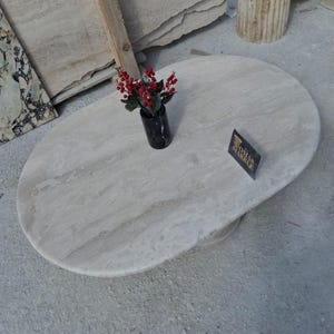 May include: A white oval marble table top with a light brown veining pattern. A small vase with red flowers sits on the table top. A sign with the text "Roma Marble" is on the table top.