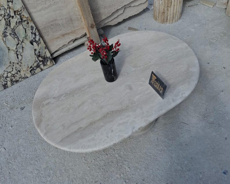 May include: A white oval marble table top with a light brown veining pattern. A small vase with red flowers sits on the table top. A sign with the text "Roma Marble" is on the table top.