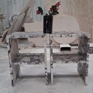 May include: Two matching side tables crafted from white and purple veined marble, each featuring a rectangular frame with a single middle shelf. A black marble vase containing a small branch with red berries sits centered on the top surface.