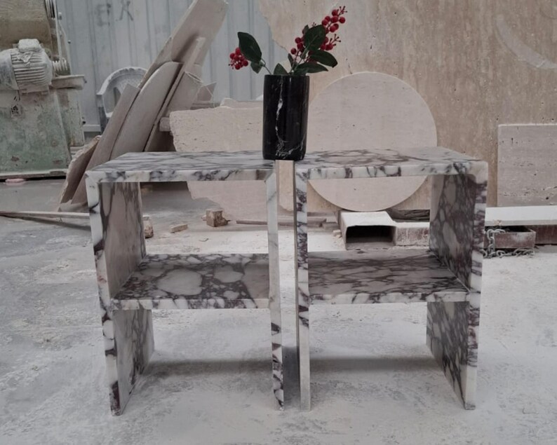 May include: Two matching side tables crafted from white and purple veined marble, each featuring a rectangular frame with a single middle shelf. A black marble vase containing a small branch with red berries sits centered on the top surface.